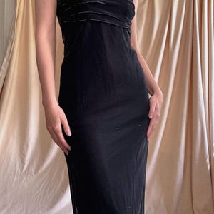 May include: A floor-length black dress featuring thin spaghetti straps and a straight neckline. The bodice is detailed with horizontal pleated fabric panels, while the skirt flows into a simple, unadorned silhouette that reaches the ankles.