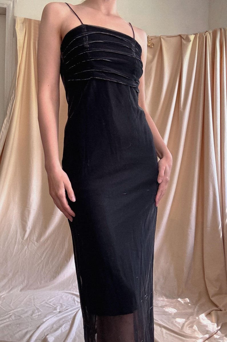 May include: A floor-length black dress featuring thin spaghetti straps and a straight neckline. The bodice is detailed with horizontal pleated fabric panels, while the skirt flows into a simple, unadorned silhouette that reaches the ankles.