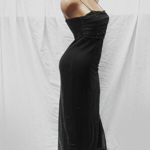 May include: A floor-length black dress featuring thin spaghetti straps and a straight neckline. The bodice includes horizontal pleated detailing, while the skirt portion is crafted from a sheer overlay fabric with subtle, scattered metallic accents.
