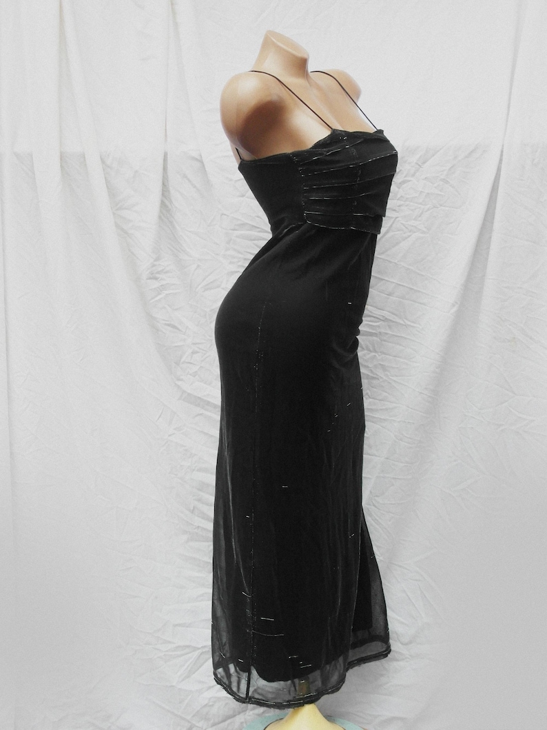May include: A floor-length black dress featuring thin spaghetti straps and a straight neckline. The bodice includes horizontal pleated detailing, while the skirt portion is crafted from a sheer overlay fabric with subtle, scattered metallic accents.