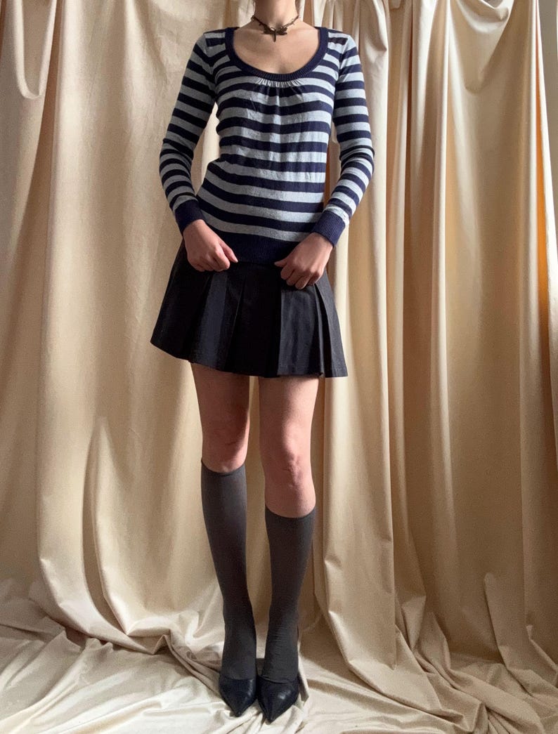 Puede incluir: Un su&eacute;ter de manga larga a rayas azul marino y gris con cuello redondo se combina con una minifalda plisada negra. El conjunto se completa con calcetines hasta la rodilla grises y tacones negros de punta. La ropa se muestra sobre un fondo neutro.