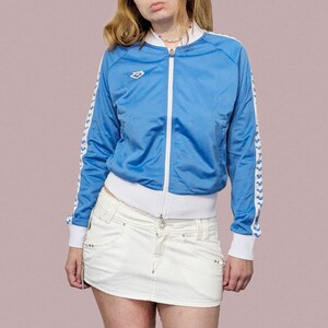May include: A light blue zip-up track jacket with white accents on the collar, cuffs, and hem. The jacket features white stripes with a repeating pattern on the sleeves. The brand logo is visible on the chest. The jacket is paired with a white denim mini skirt.