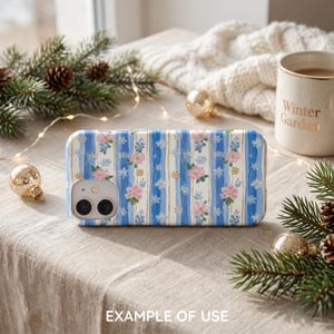 May include: A rectangular smartphone case featuring a vertical pattern of blue brushstroke stripes, pink flowers, and white snowflakes. The case has a cutout for a dual-lens camera system on the upper left corner and a smooth, glossy finish.