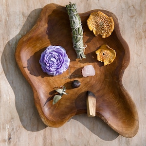 May include: A wooden bowl containing a selection of items, including a purple flower, a sage smudge stick, a pink crystal, a small piece of wood, and two dried orange peels.