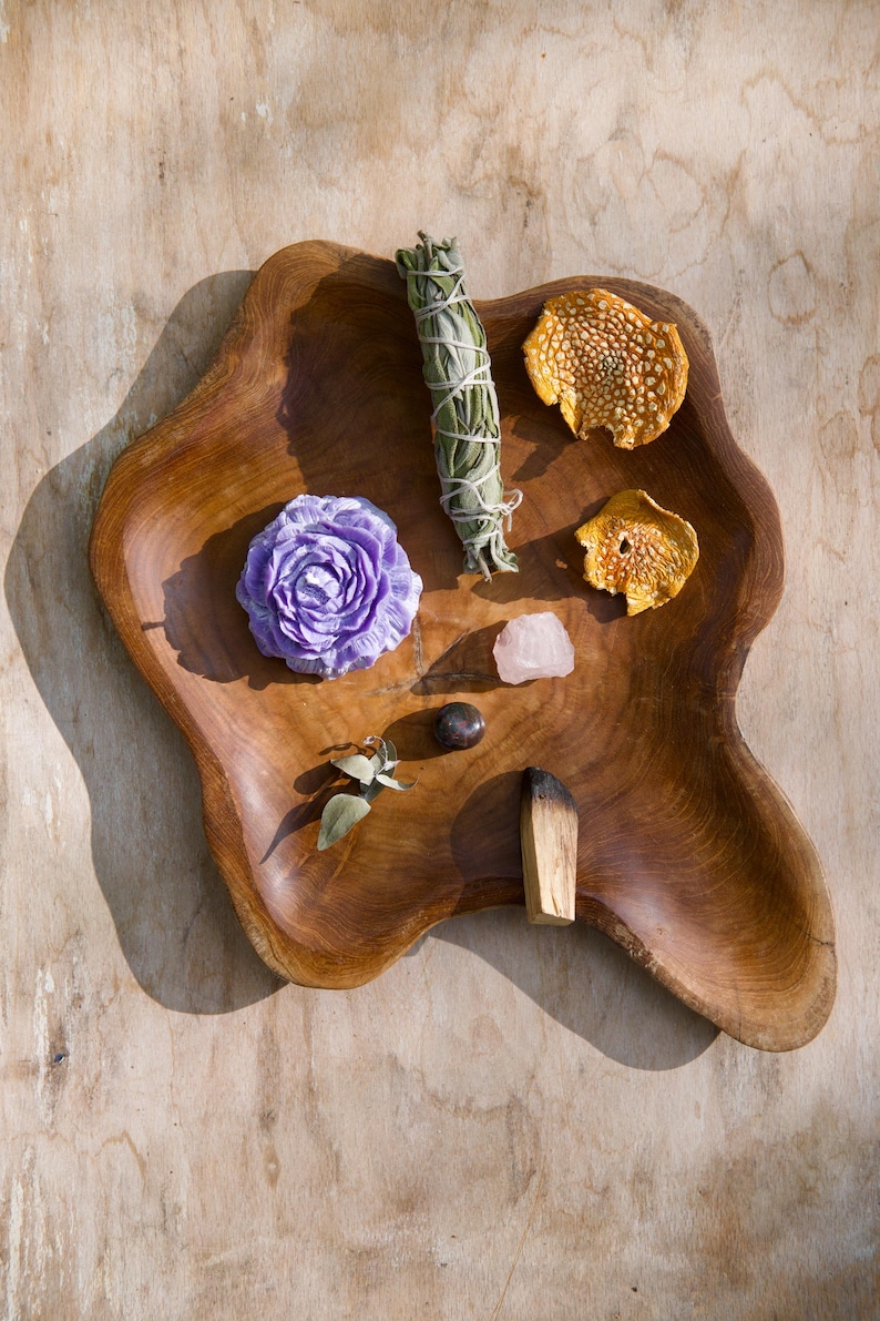 May include: A wooden bowl containing a selection of items, including a purple flower, a sage smudge stick, a pink crystal, a small piece of wood, and two dried orange peels.