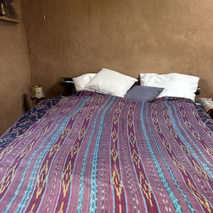 Könnte beinhalten: Ein Bett mit einer lila, blauen und gelb gestreiften Decke. Die Decke ist über das Bett drapiert, das weiße Kissen und ein dunkles Kopfteil hat. Der Raum hat braune Wände.