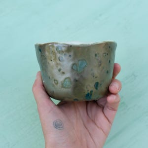 Puede incluir: Una taza pequeña de cerámica moteada con un esmalte marrón claro y motas azules y verdes. La taza está siendo sostenida en una mano.