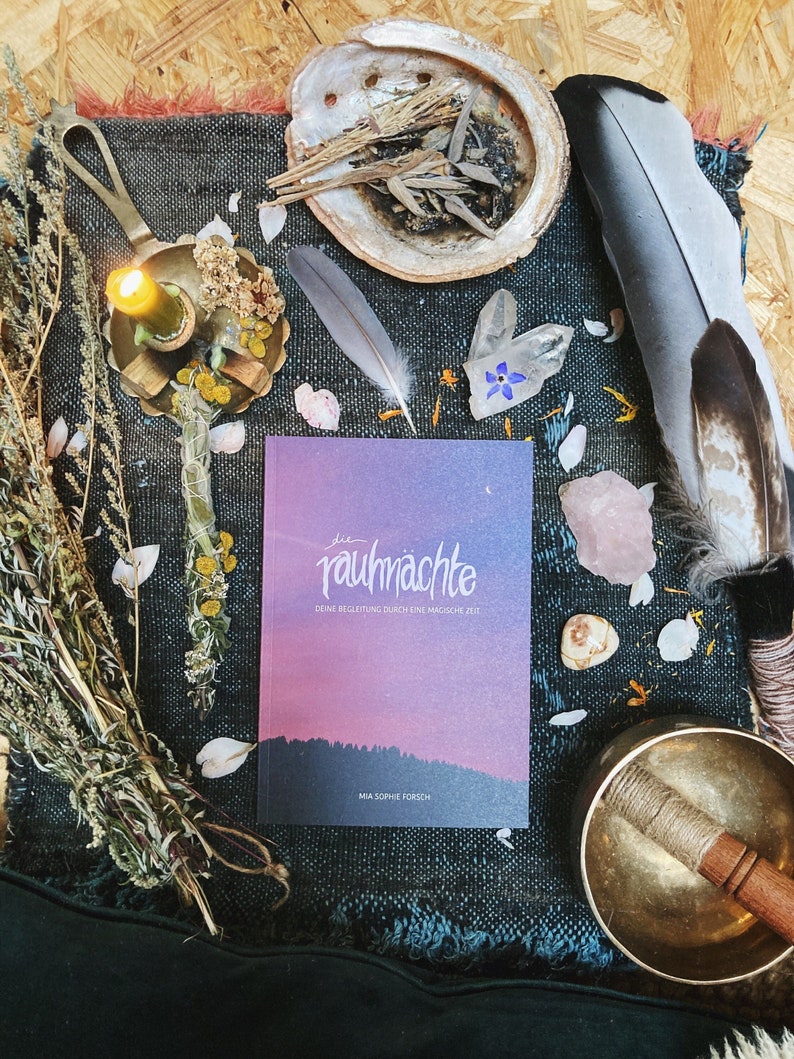 May include: A book titled "Die Rauchn&auml;chte: Deine Begleitung durch eine magische Zeit" by Mia Sophie F&ouml;rch, with a purple and pink gradient cover, surrounded by various objects including a candle, feathers, crystals, and a singing bowl.