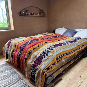 May include: A colorful woven blanket with a geometric pattern in shades of yellow, pink, black, and gray covers a bed made of bamboo logs. The blanket has a fringed edge and is placed on a wooden floor. A patterned rug is on the floor.