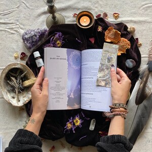 May include: An open book with the title "Balance and Rhythm" on a purple velvet surface. The book is surrounded by candles, crystals, feathers, and other objects. The book is open to a page with a mandala design and text in German.