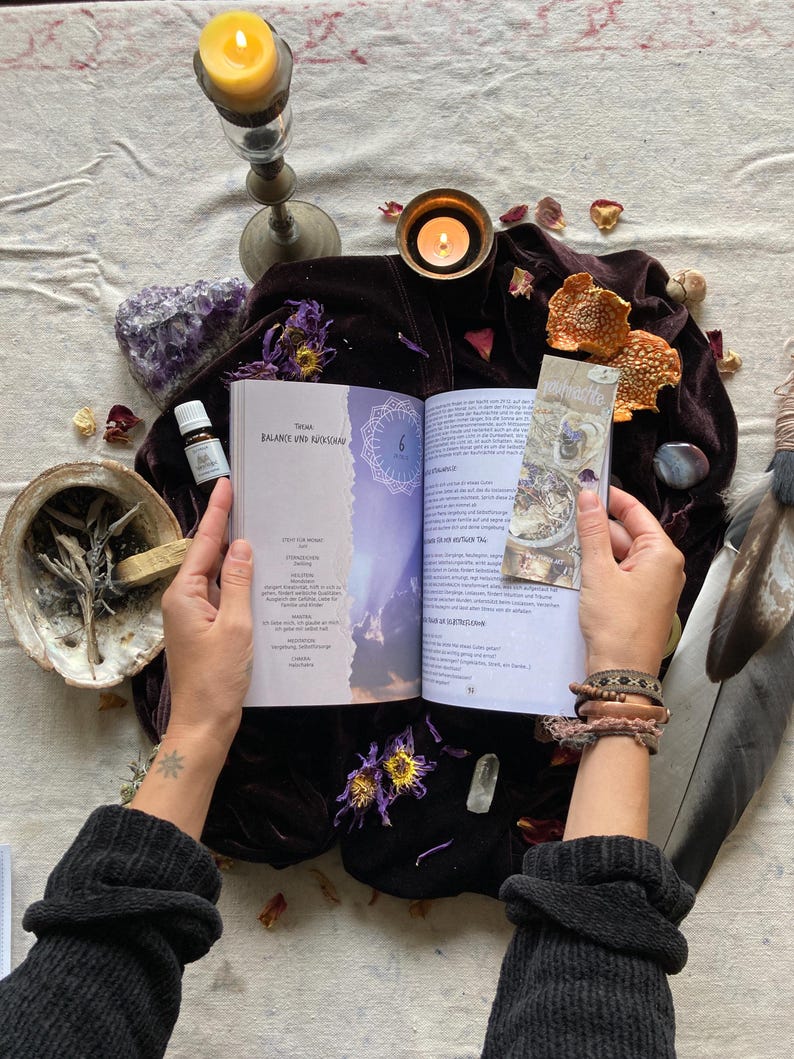 May include: An open book with the title "Balance and Rhythm" on a purple velvet surface. The book is surrounded by candles, crystals, feathers, and other objects. The book is open to a page with a mandala design and text in German.