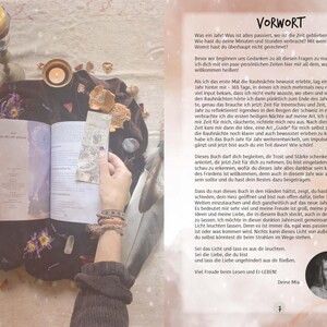 May include: A book with a white cover and the title "Year of the Moon" in black text. The book is open and a hand is holding the pages. The background is a soft, light pink with a variety of objects, including candles, flowers, and crystals.
