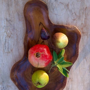 May include: A wooden serving tray with a natural wood grain finish holds a red pomegranate, a green pear, two green figs, and a sprig of green sage.