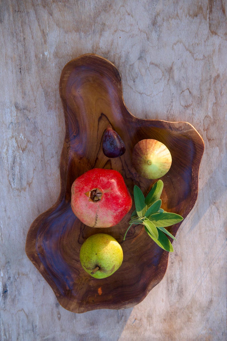 May include: A wooden serving tray with a natural wood grain finish holds a red pomegranate, a green pear, two green figs, and a sprig of green sage.