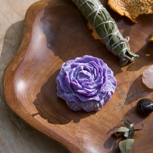 May include: A purple flower-shaped candle sits on a wooden tray with a bundle of sage, a dried orange peel, a small piece of rose quartz, and a black stone.