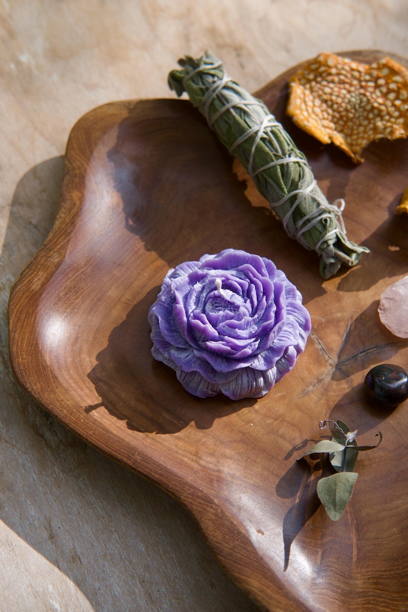 May include: A purple flower-shaped candle sits on a wooden tray with a bundle of sage, a dried orange peel, a small piece of rose quartz, and a black stone.