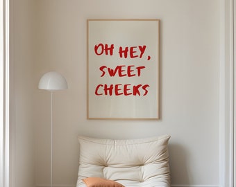 May include: A white, overstuffed chair with a red pillow sits in a room with white walls and wooden floors. A framed print with the text "Oh hey, sweet cheeks" hangs on the wall behind the chair.
