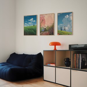 May include: Three framed art prints of landscapes with couples walking in the distance. The prints are hung on a white wall above a dark blue couch and a white shelf with a book titled "ISLANDS" and a black vase.