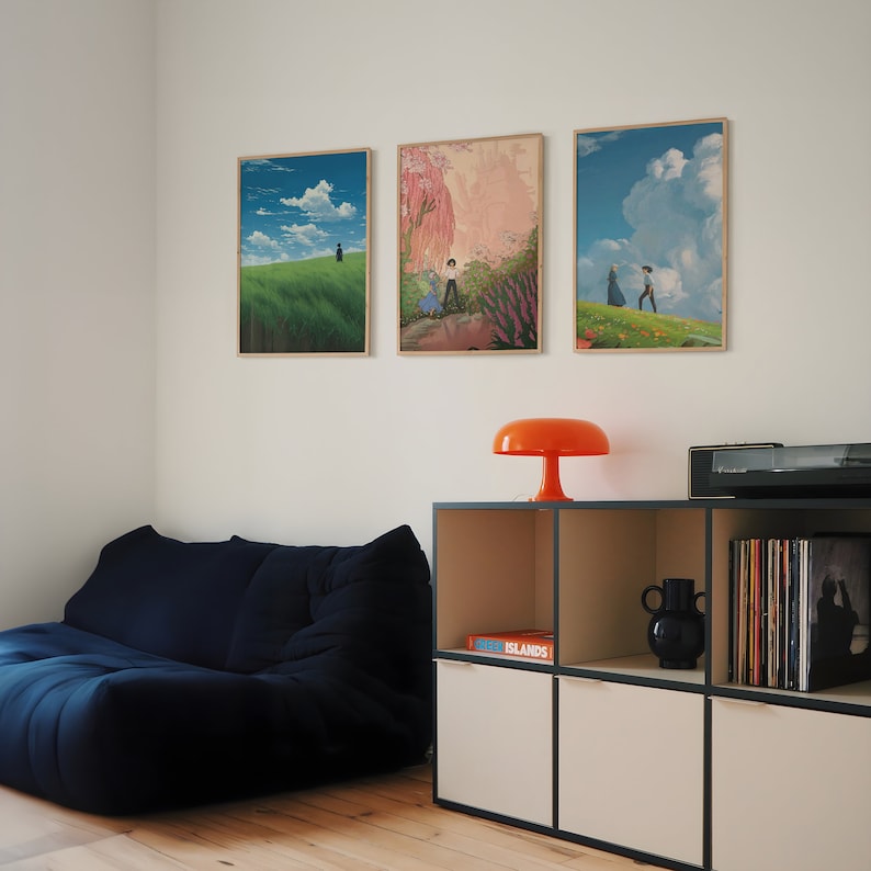 May include: Three framed art prints of landscapes with couples walking in the distance. The prints are hung on a white wall above a dark blue couch and a white shelf with a book titled "ISLANDS" and a black vase.