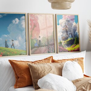 May include: Three framed prints of anime artwork. The first print features a blue sky with white clouds and two figures walking on a green hill. The second print shows a pink and green landscape with a figure in a blue dress and a figure in a white shirt. The third print features a floating house in a blue sky with white clouds and a green landscape.