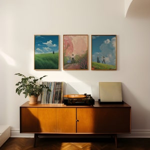 May include: Three framed art prints with a minimalist aesthetic, featuring landscapes with figures in the distance. The prints are hung on a white wall above a mid-century modern wooden cabinet with a record player and vinyl records.