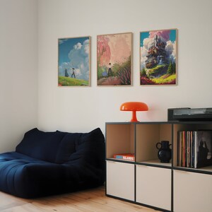 May include: Three framed art prints on a white wall above a dark blue couch and a white and black shelving unit. The prints feature colorful illustrations of a couple walking in a field, a woman walking in a field, and a flying house.