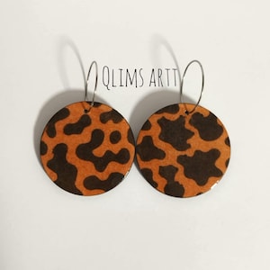 Puede incluir: Un par de pendientes redondos con un estampado de animal marrón y naranja. Los pendientes tienen aros plateados. El texto "QLIMS ARTT" está encima de los pendientes.