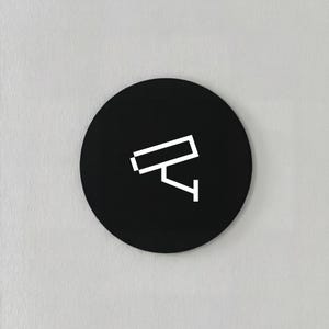 May include: A black, circular sign featuring a white, minimalist security camera icon. The camera is depicted with simple geometric shapes, suggesting surveillance or monitoring. The sign is mounted on a light gray wall.