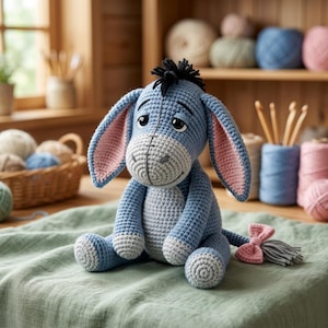 Pode incluir: Um brinquedo de pelúcia de burro de crochê azul e cinza feito à mão, com o interior das orelhas rosa e uma crina preta. A pelúcia está sentada em um pano verde, com um laço rosa e uma borla cinza ao lado. O fundo inclui fios e agulhas de crochê.