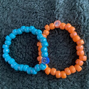 Könnte beinhalten: Zwei Armbänder aus Perlen. Eines ist hellblau, das andere orange. Das blaue Armband hat einen kleinen blau-weißen Anhänger. Das orangefarbene Armband hat einen kleinen Anhänger mit einem Eulenmuster. Die Armbänder bestehen aus kleinen, abgerundeten Perlen.