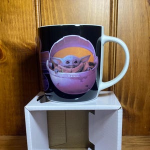 K&ouml;nnte beinhalten: Schwarze Keramik-Tasse mit einer Grafik von Grogu, auch bekannt als Baby Yoda, in einer rosa Trage. Die Tasse hat einen wei&szlig;en Henkel und steht auf einem wei&szlig;en Pappkarton. Der Hintergrund ist eine Holzoberfl&auml;che.