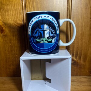 K&ouml;nnte beinhalten: Eine blaue Keramiktasse mit dem Text "READY FOR ADVENTURE", der ein Bild eines Mandalorianer-Helms und Grogu umgibt. Die Tasse hat einen wei&szlig;en Henkel und steht auf einem wei&szlig;en Karton.