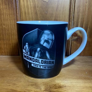 May include: Black ceramic mug with a white handle and interior. The mug features a black and white image of The Mandalorian with the text "THE MANDALORIAN THIS IS THE WAY" in white.
