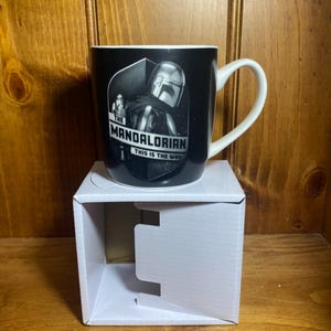 May include: Black ceramic mug featuring The Mandalorian. The mug has a white handle and rim. The front of the mug displays an image of the Mandalorian and the text "THE MANDALORIAN THIS IS THE WAY". The mug is sitting on a white cardboard box.