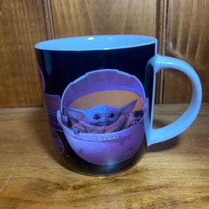 K&ouml;nnte beinhalten: Wei&szlig;er Keramikbecher mit einer Grafik von Grogu, auch bekannt als Baby Yoda, in seiner schwebenden Wiege. Der Becher hat einen schwarzen Hintergrund mit hellblauem Innenraum und Griff. Das Bild stammt aus der Star Wars-Serie The Mandalorian.