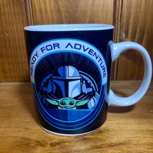K&ouml;nnte beinhalten: Eine wei&szlig;e Keramiktasse mit blauem &Auml;u&szlig;eren, die den Text "READY FOR ADVENTURE" um ein Bild eines Mandalorianer-Helms und Grogu tr&auml;gt. Die Tasse hat einen wei&szlig;en Henkel und ein wei&szlig;es Inneres.