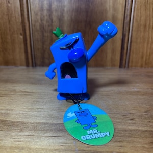 Puede incluir: Una figura azul de Mr. Grumpy con un brote verde en la parte superior y un brazo levantado en un puño. La figura tiene una cuerda negra unida a una etiqueta con las palabras "MR. GRUMPY" y una ilustración del personaje.