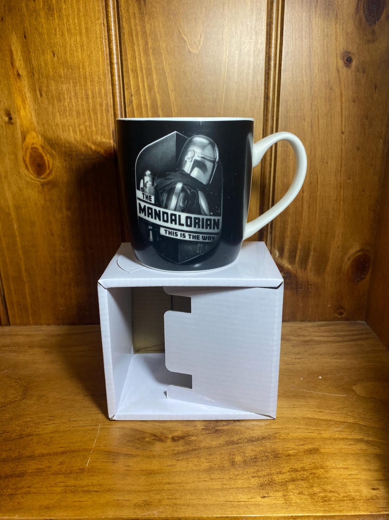 May include: Black ceramic mug featuring The Mandalorian. The mug has a white handle and rim. The front of the mug displays an image of the Mandalorian and the text "THE MANDALORIAN THIS IS THE WAY". The mug is sitting on a white cardboard box.