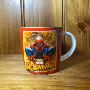 Puede incluir: Taza de cer&aacute;mica roja con asa blanca, con Spider-Man en cuclillas sobre un fondo de telara&ntilde;a amarilla. Tambi&eacute;n se ve el logotipo de Marvel Spider-Man.