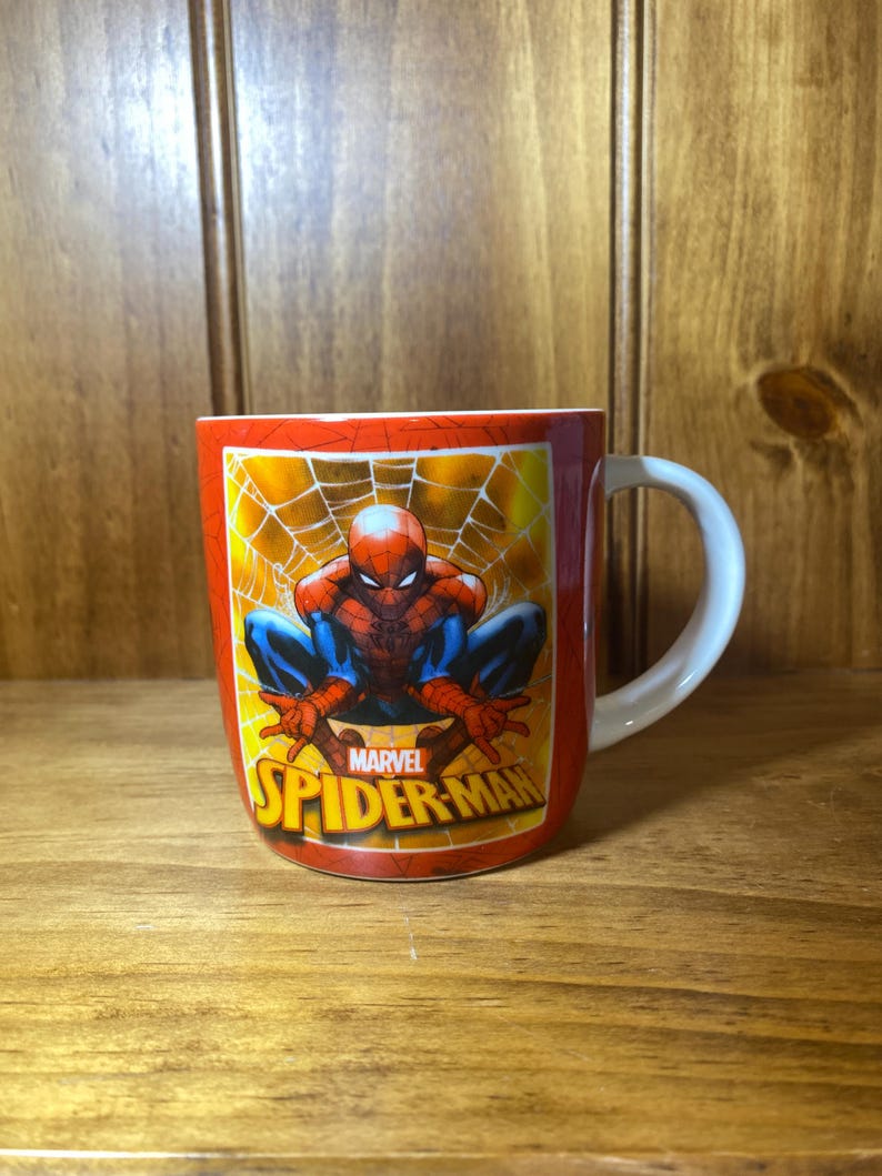 Puede incluir: Taza de cer&aacute;mica roja con asa blanca, con Spider-Man en cuclillas sobre un fondo de telara&ntilde;a amarilla. Tambi&eacute;n se ve el logotipo de Marvel Spider-Man.