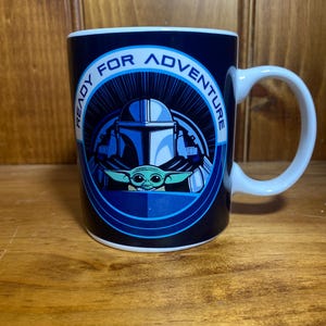 K&ouml;nnte beinhalten: Wei&szlig;er Keramikbecher mit einer blau-wei&szlig;en Grafik mit The Mandalorian und Grogu. Der Becher tr&auml;gt die Worte "READY FOR ADVENTURE" in einem kreisf&ouml;rmigen Design um die Charaktere. Der Becher hat einen wei&szlig;en Griff.
