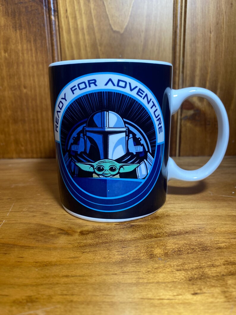 K&ouml;nnte beinhalten: Wei&szlig;er Keramikbecher mit einer blau-wei&szlig;en Grafik mit The Mandalorian und Grogu. Der Becher tr&auml;gt die Worte "READY FOR ADVENTURE" in einem kreisf&ouml;rmigen Design um die Charaktere. Der Becher hat einen wei&szlig;en Griff.