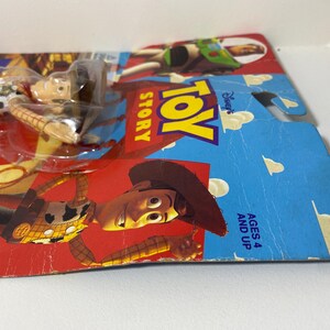 以下が含まれることがあります： ウッディのトイストーリーアクションフィギュアのパッケージ。ウッディは赤いバンダナと茶色の帽子をかぶったカウボーイ人形です。パッケージは青と白で、「Disney's Toy Story」と「Ages 4 and Up」の文字が書かれています。