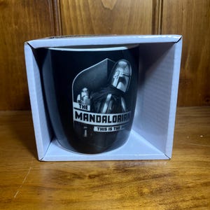 May include: Black ceramic mug featuring a grayscale image of The Mandalorian character. The mug is in a white box and has the text "THE MANDALORIAN THIS IS THE WAY". The mug is cylindrical and appears to be new.