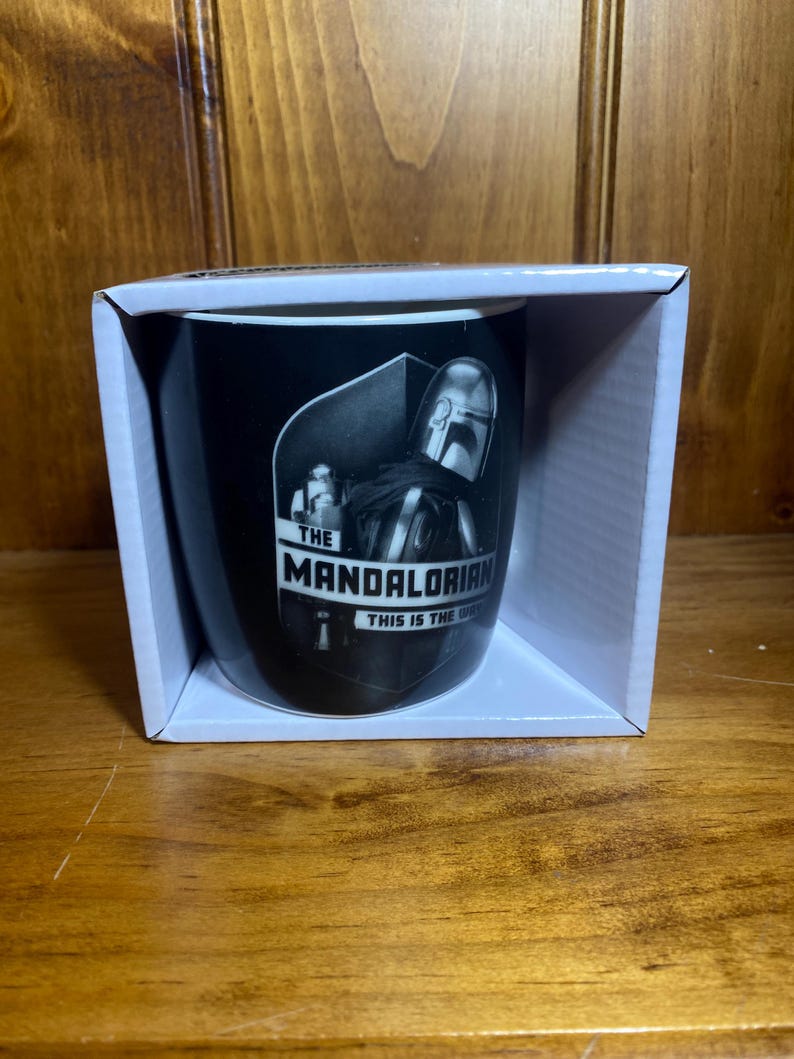 May include: Black ceramic mug featuring a grayscale image of The Mandalorian character. The mug is in a white box and has the text "THE MANDALORIAN THIS IS THE WAY". The mug is cylindrical and appears to be new.