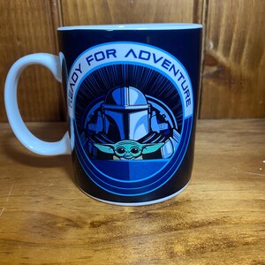 K&ouml;nnte beinhalten: Eine wei&szlig;e Keramiktasse mit einem blau-schwarzen Design mit einem Mandalorianer-Helm und Grogu. Auf der Tasse steht in einem kreisf&ouml;rmigen Muster "READY FOR ADVENTURE". Die Tasse hat einen wei&szlig;en Henkel.