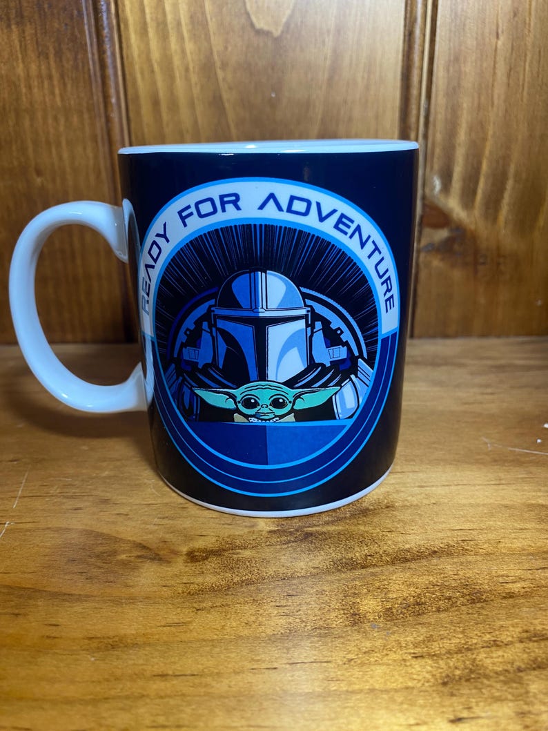 K&ouml;nnte beinhalten: Eine wei&szlig;e Keramiktasse mit einem blau-schwarzen Design mit einem Mandalorianer-Helm und Grogu. Auf der Tasse steht in einem kreisf&ouml;rmigen Muster "READY FOR ADVENTURE". Die Tasse hat einen wei&szlig;en Henkel.
