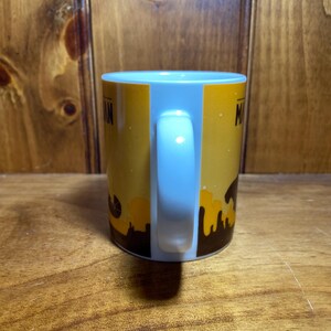 Puede incluir: Una taza de cer&aacute;mica blanca con un dise&ntilde;o gr&aacute;fico en amarillo y marr&oacute;n que presenta la silueta de un horizonte urbano en la base. La taza tiene un asa curva en el lateral y texto parcial visible sobre el fondo amarillo, mostrando las letras M y A.