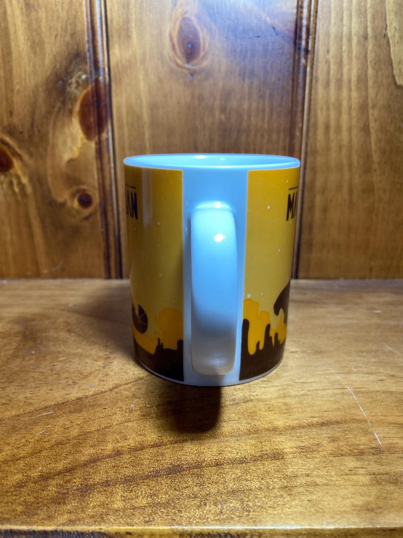 Puede incluir: Una taza de cer&aacute;mica blanca con un dise&ntilde;o gr&aacute;fico en amarillo y marr&oacute;n que presenta la silueta de un horizonte urbano en la base. La taza tiene un asa curva en el lateral y texto parcial visible sobre el fondo amarillo, mostrando las letras M y A.