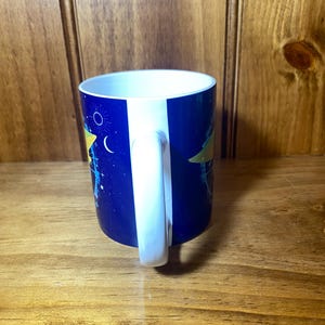 May include: A ceramic mug with a white handle and interior. The exterior features a dark blue background with a celestial design including stars, a crescent moon, and a yellow shape. The mug is on a wooden surface.